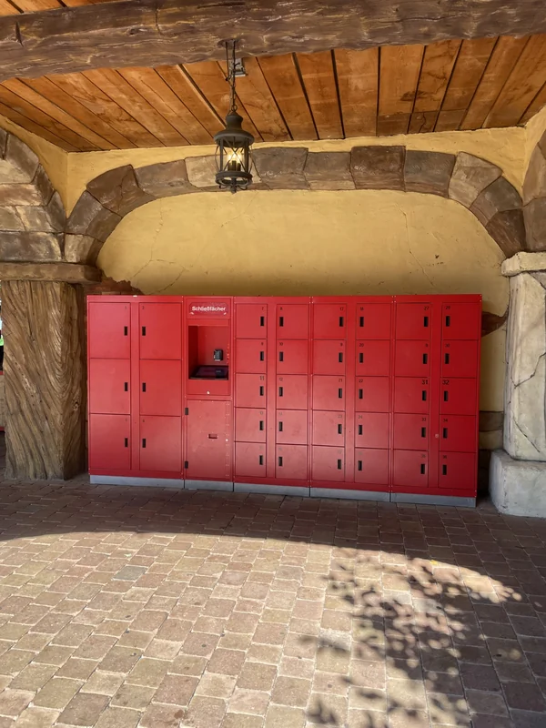 Red self-service lockers with a payment kiosk beneath a timber and stone shelter at an earlier theme-park installation.