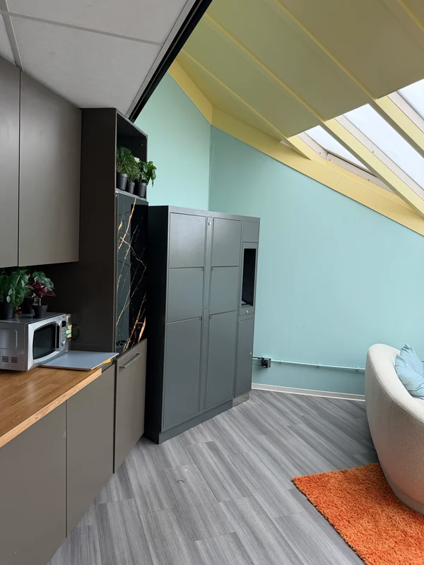 Compact grey locker bank beside a kitchenette and seating in a hotel interior.