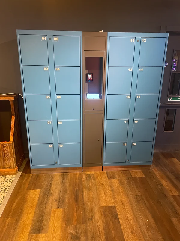 Blue-grey electronic lockers arranged on either side of a central payment kiosk in a competitive socialising venue.