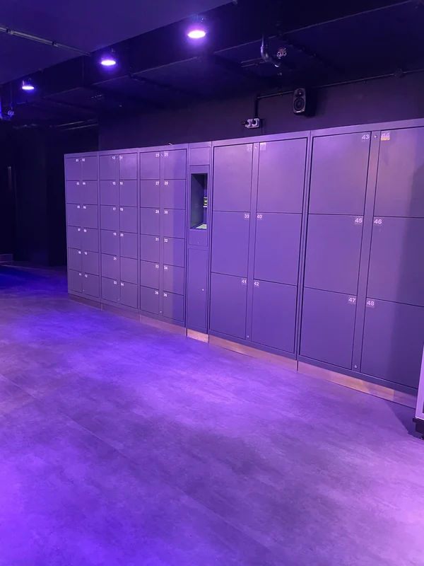 Grey self-service lockers with small and large compartments under purple lighting at a competitive socialising venue.