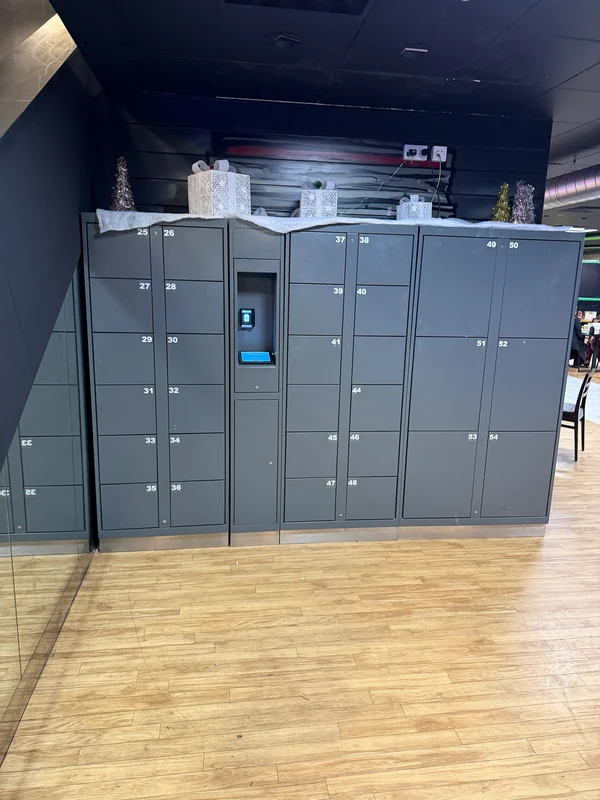 Grey electronic lockers with different compartment sizes and a central kiosk beside a mirrored wall.