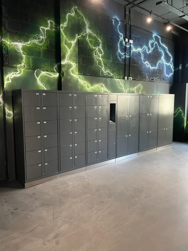 Grey self-service locker bank with small and larger compartments beneath a lightning mural in a family entertainment centre.