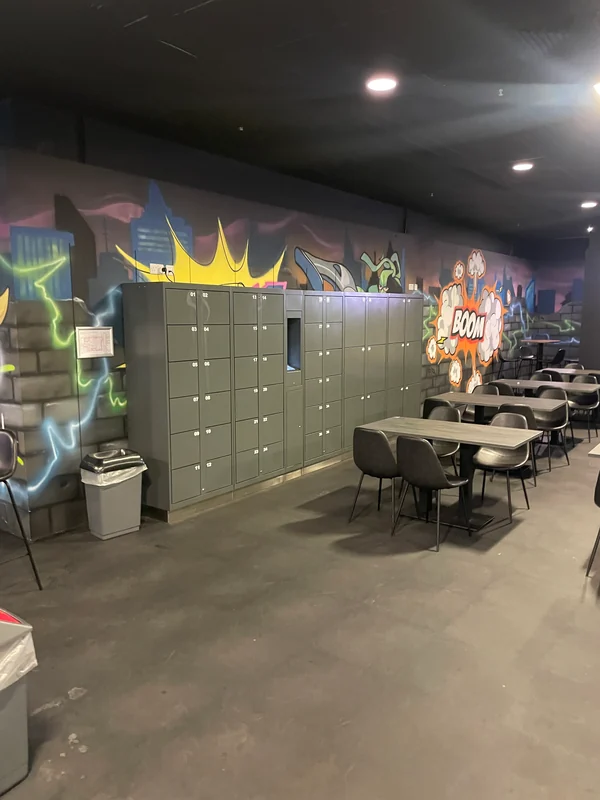 Grey self-service lockers beside tables and chairs in a family entertainment centre, with a comic-style wall mural behind.