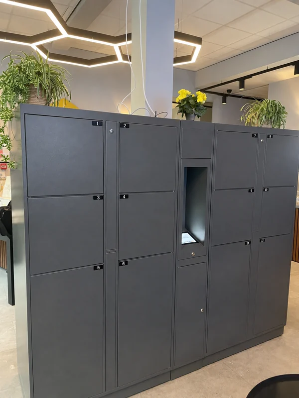 Grey hotel lockers with a central touchscreen recess beside plants and geometric ceiling lights.