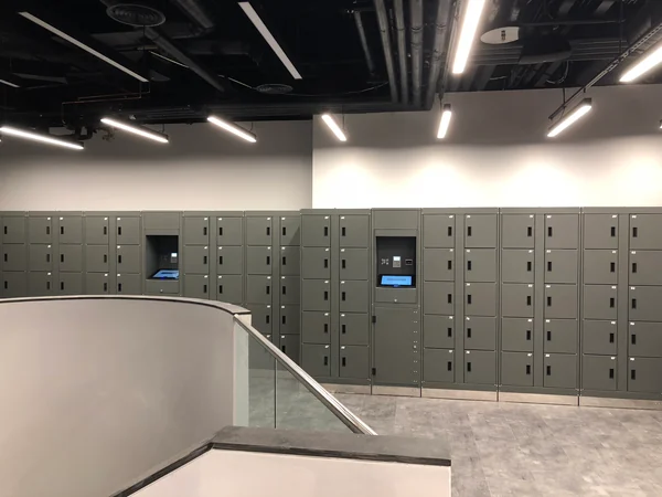 Grey self-service lockers with two touchscreen kiosks beside a stairwell at an earlier museum installation.
